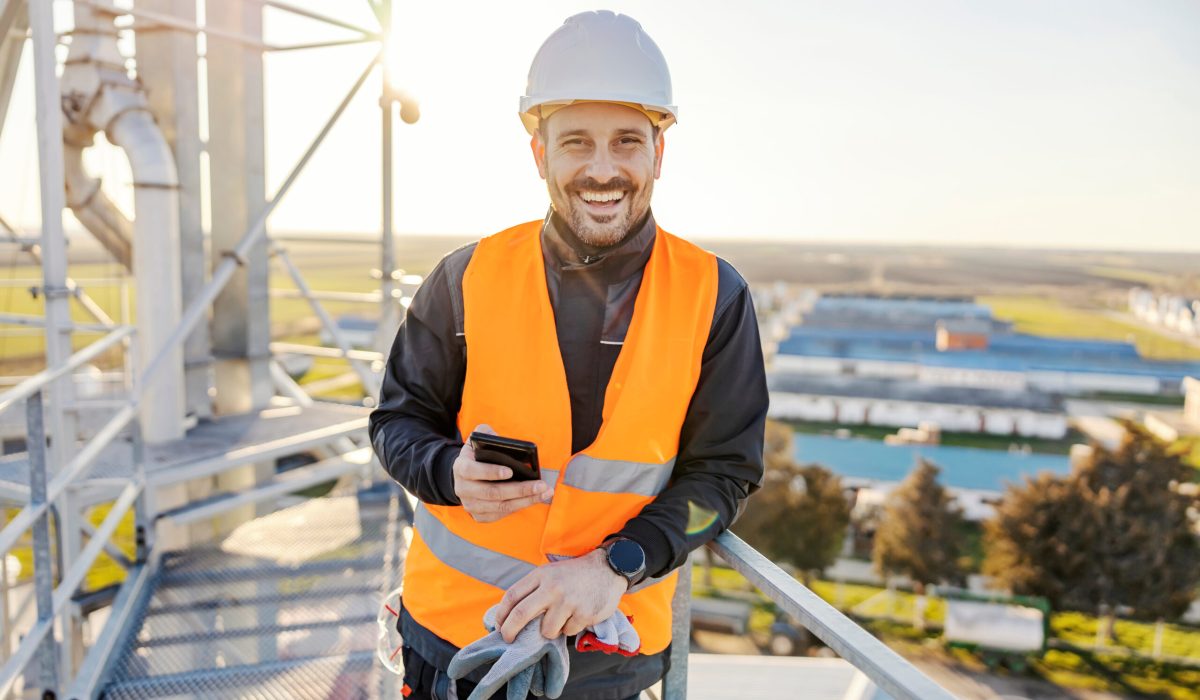 A happy industry worker holding phone on height on metal construction and smiling.